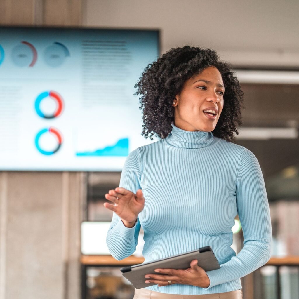 Woman holding tablet and standing in front of large television screen with pie charts on it.