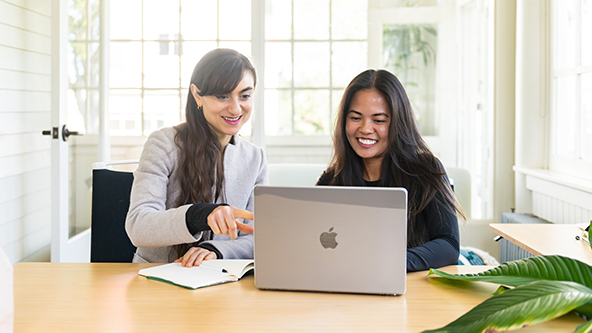 Two females working together at a table. Looking at a laptop. Both are smiling.